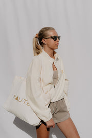 Woman wearing a white jacket and sunglasses, holding a tote bag with 'HAUTE' branding, against a plain background.