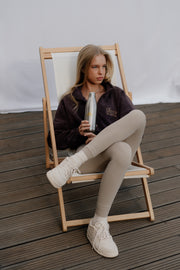 Woman sitting on a wooden chair holding a bottle, wearing a dark jacket and light pants.