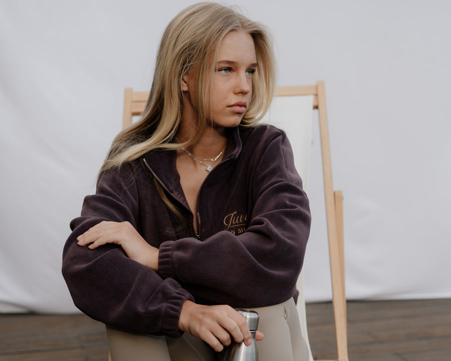 Woman sitting on a wooden chair holding a thermos, wearing a dark jacket and light pants.
