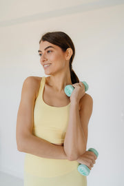 Woman in a yellow sports bra holding green dumbbells against a white background