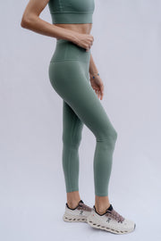 Woman wearing green athletic leggings and a sports bra on a light gray background