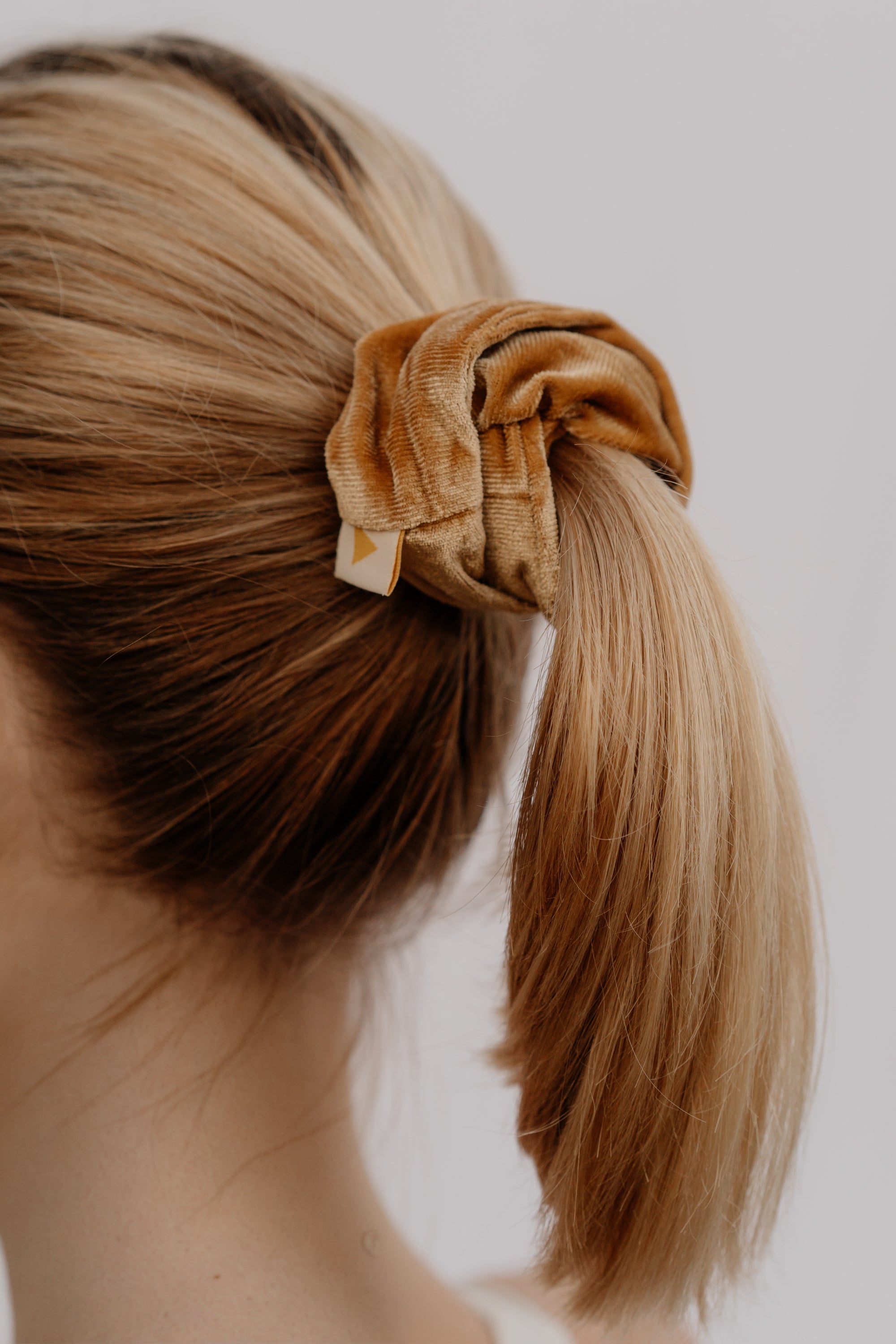 Blonde hair tied into a ponytail with a golden scrunchie against a neutral background