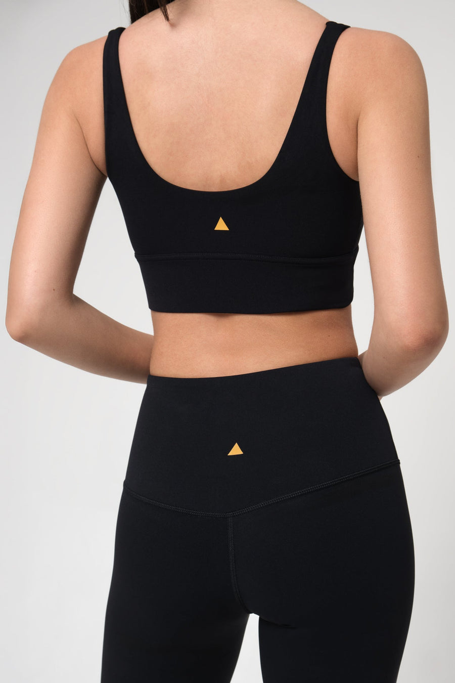 Woman wearing a black sports bra and leggings with a small yellow triangle logo on a light gray background