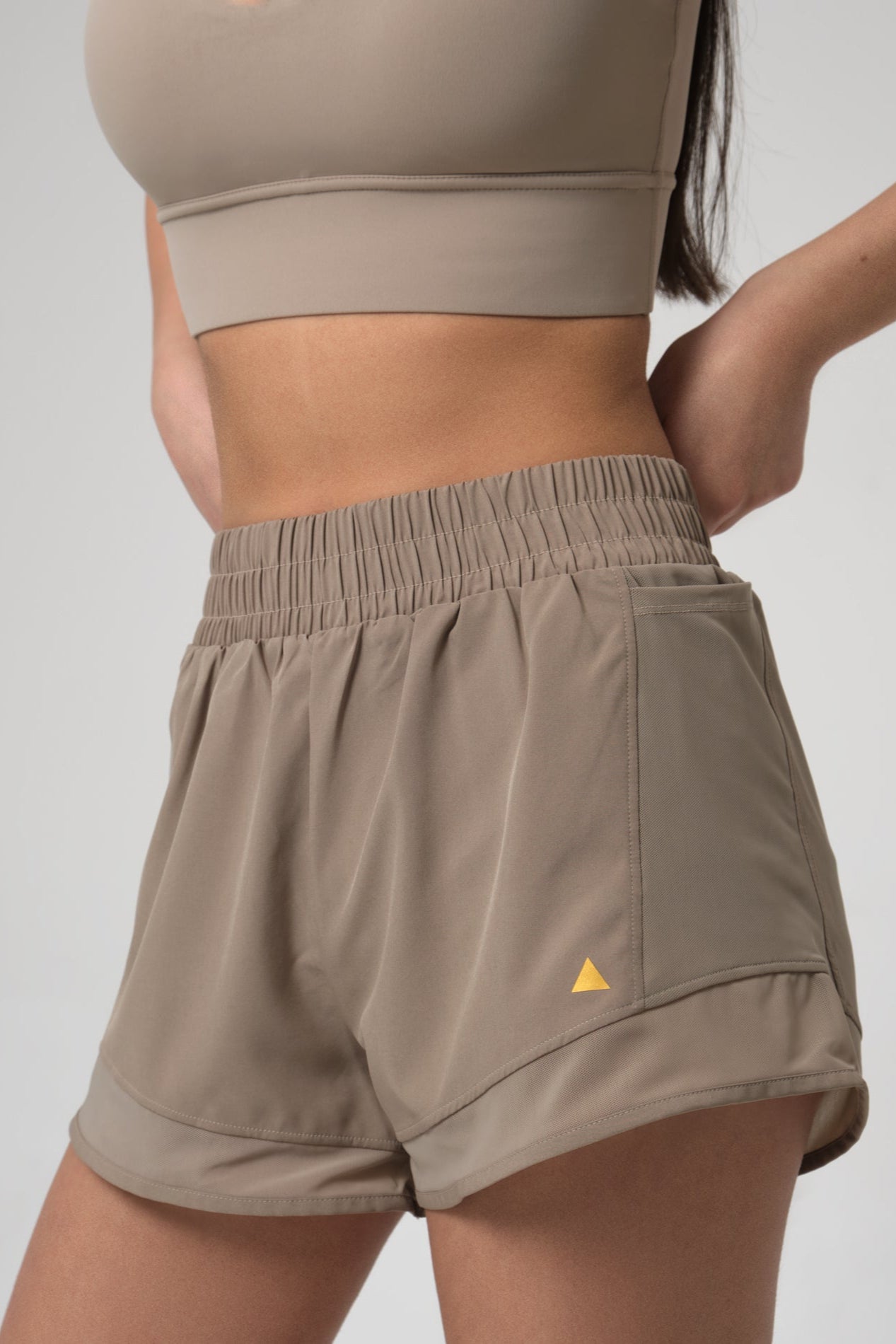 Woman wearing beige athletic shorts with a small yellow triangle logo on a plain background