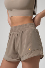 Woman wearing beige athletic shorts with a small yellow triangle logo on a plain background