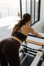 Woman exercising on a Pilates reformer machine in a bright room.