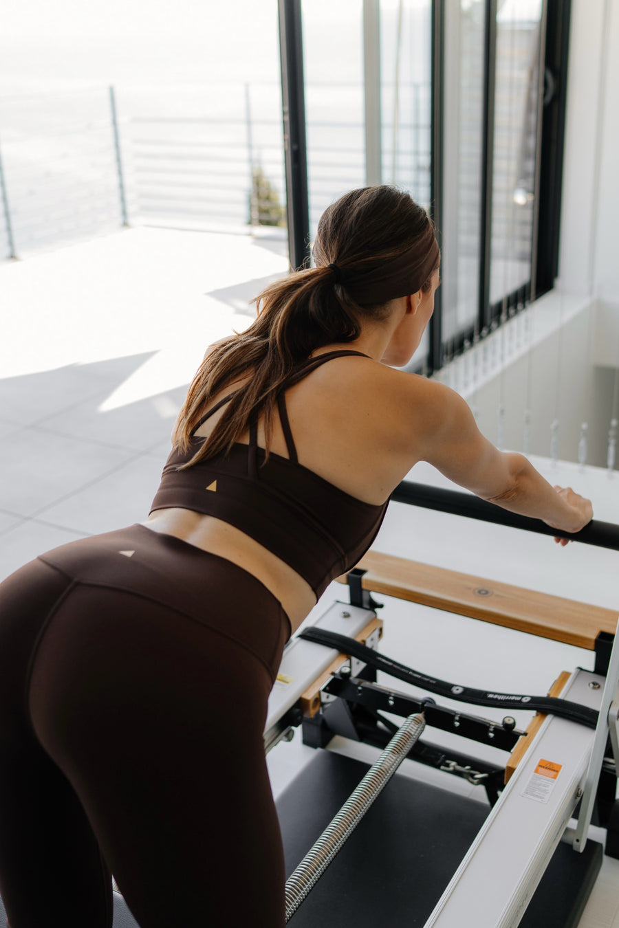 Woman exercising on a Pilates reformer machine in a bright room.