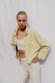 Woman wearing a light green jacket and white leggings against a plain background