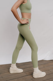 Woman wearing green athletic leggings and top on a wooden floor with white background