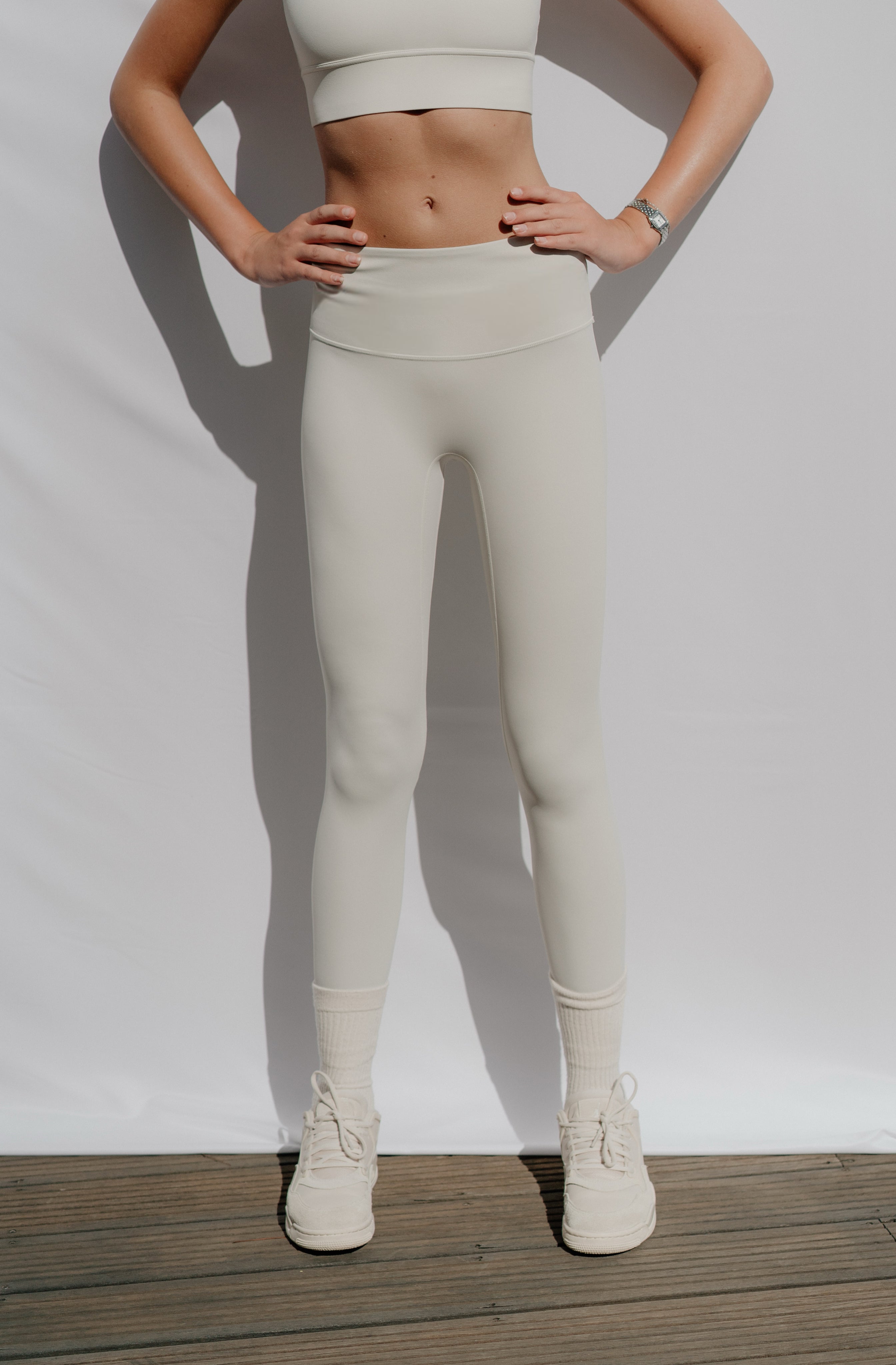 Woman wearing beige leggings and a white top on a plain background