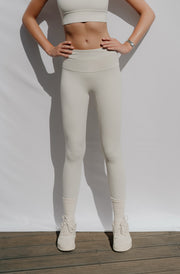 Woman wearing beige leggings and a white top on a plain background