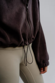 Woman wearing a dark brown jacket and beige pants against a neutral background